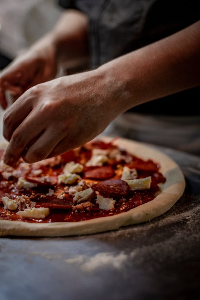 Hands of a chef skillfully preparing a pepperoni pizza with fresh ingredients in a kitchen setting.
